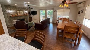 A spacious living room with a fireplace, a dining area with wooden furniture, and a stone accent wall. - Creekside Dream - Dog Friendly/Game Room/Sleeps 12 (Oakhurst)