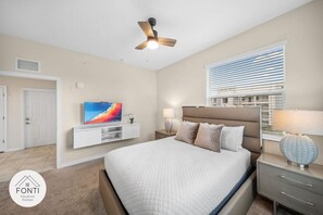 Ein Schlafzimmer mit einem Bett, einem Fernseher auf einer weißen Kommode, einem Deckenventilator, einem Fenster mit Jalousien und einem Nachttisch mit Lampe.