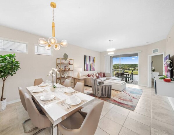 A spacious living area with a dining table set for a meal, a sofa, and a coffee table. There is a large window providing a view of the outdoors.