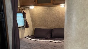 A compact RV bedroom with a bed, wooden cabinets, and a window with blinds. - 19FT RAPTOR RV (Wasilla)