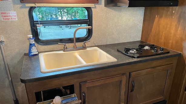 A compact kitchenette with a sink, stove, and storage cabinets. - 19FT RAPTOR RV (Wasilla)