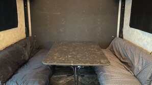 A compact RV interior with a small table and two seating areas, one with a dark cushion and the other with a patterned cushion. - 19FT RAPTOR RV (Wasilla)