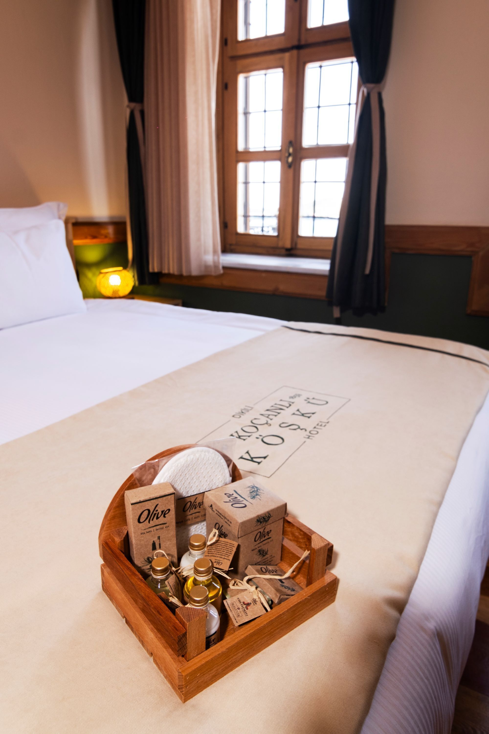 A wooden tray with olive oil bottles and boxes on a bed in a hotel room with a window and curtains.