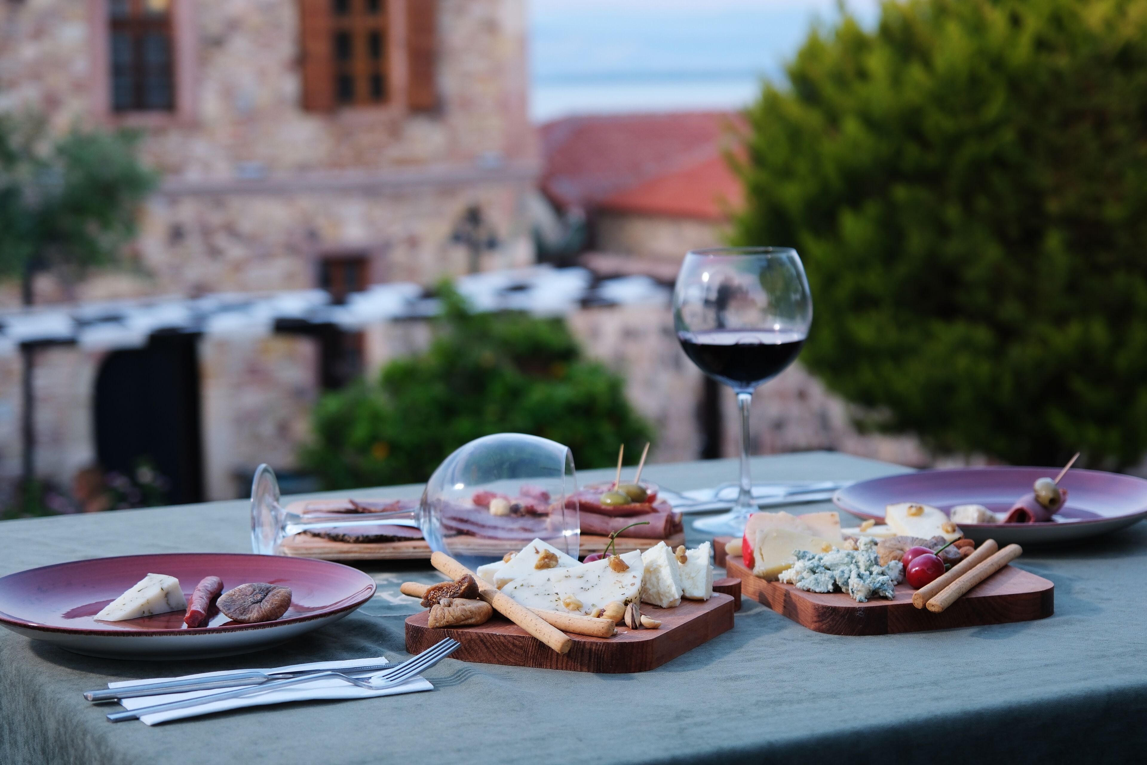 Un montaje para comer al aire libre con varios quesos y embutidos en platos, una copa de vino y un rústico edificio de piedra al fondo.