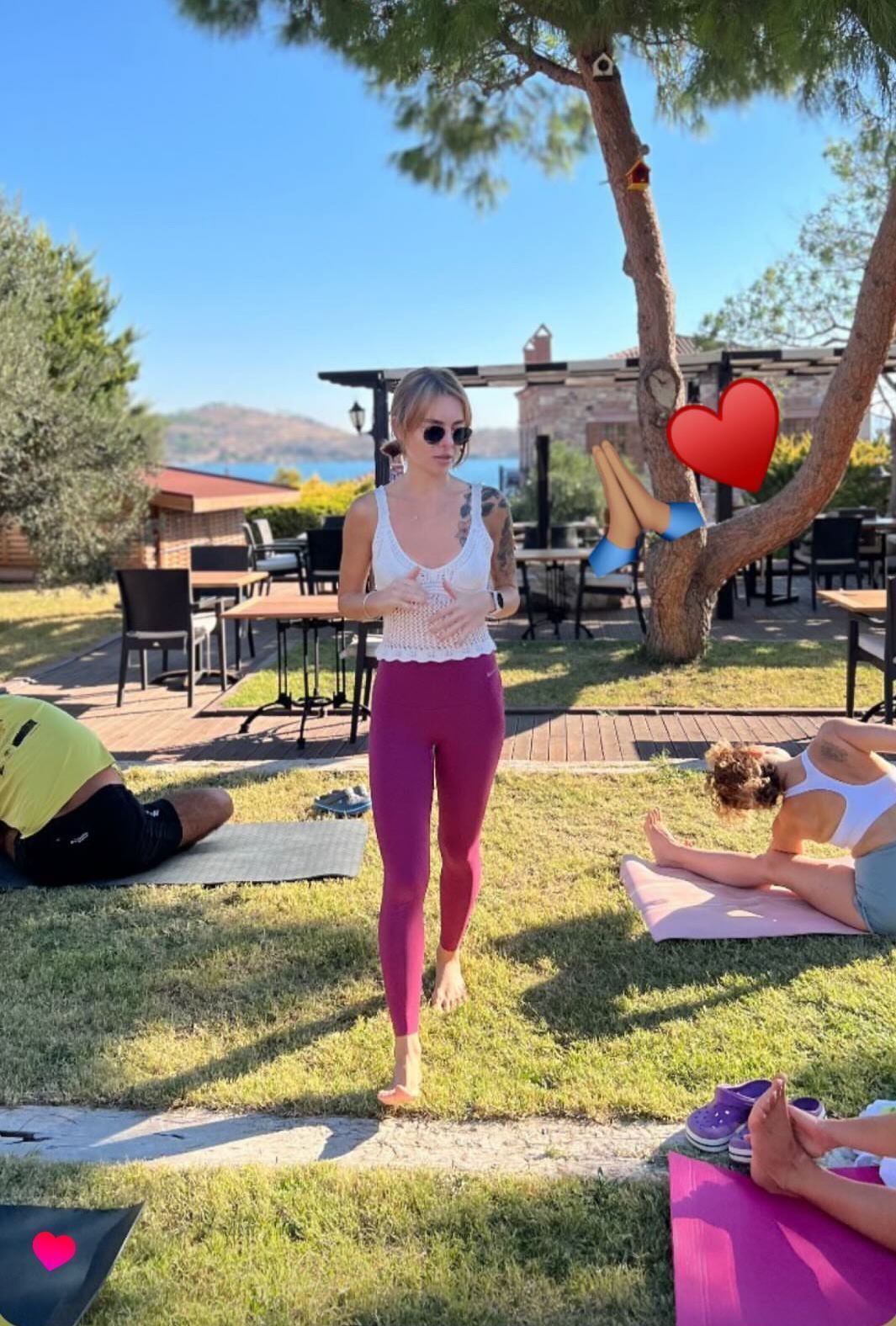 Sesión de yoga al aire libre con participantes en colchonetas, un árbol y un edificio al fondo.