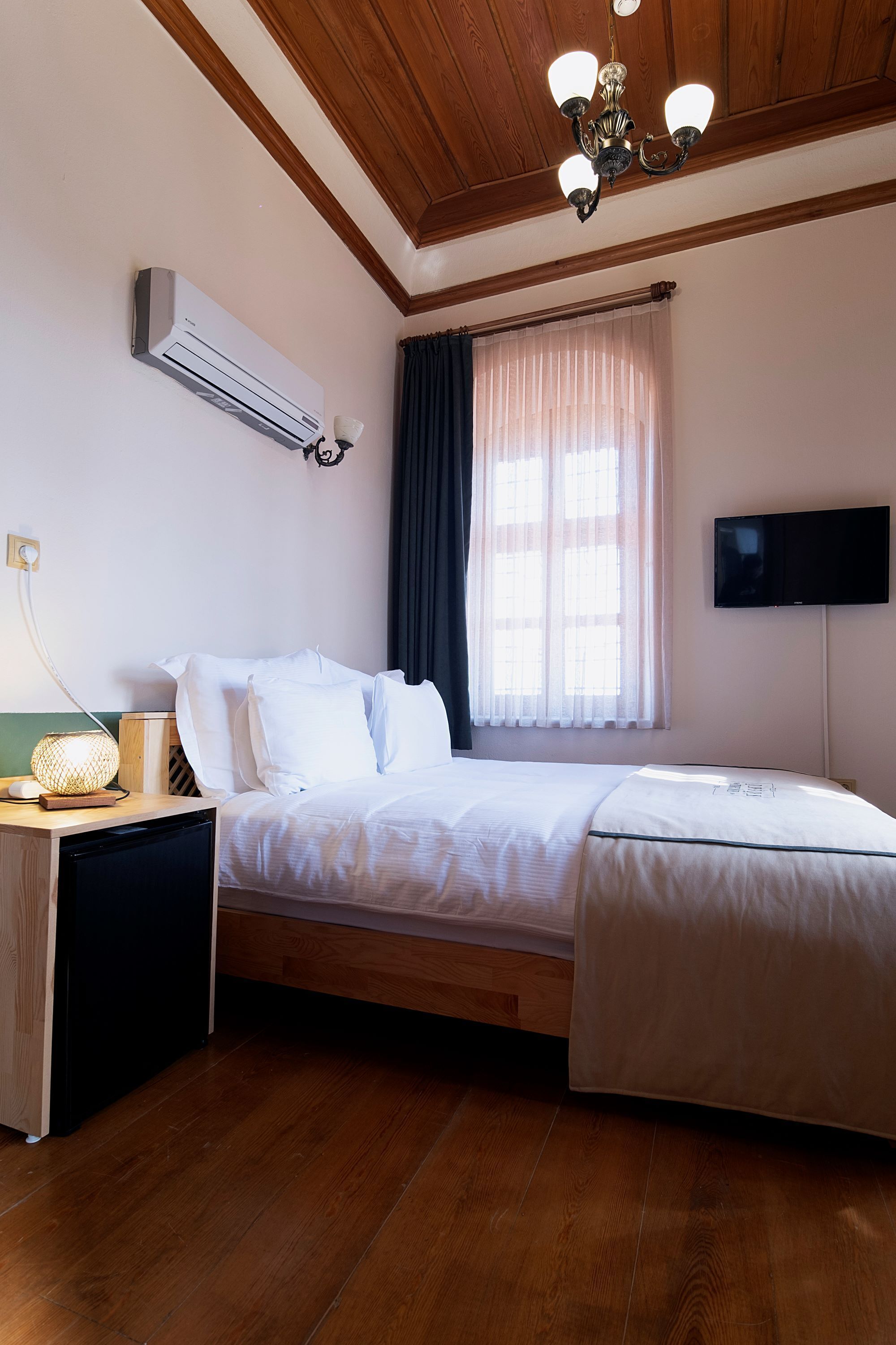 Un dormitori amb un llit, una taula de nit, una làmpada, aire condicionat, una finestra amb cortines i un televisor de pantalla plana penjat a la paret.