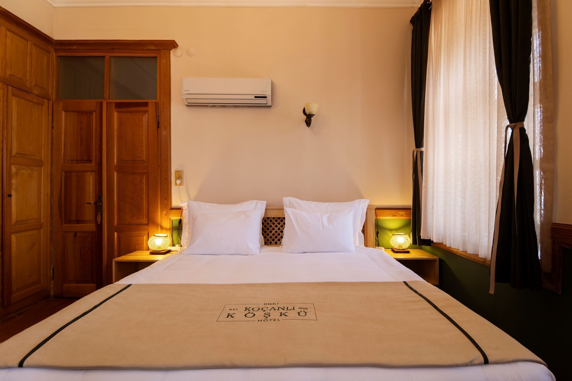 A hotel room with a bed, wooden wardrobe, air conditioning unit, and two bedside lamps.