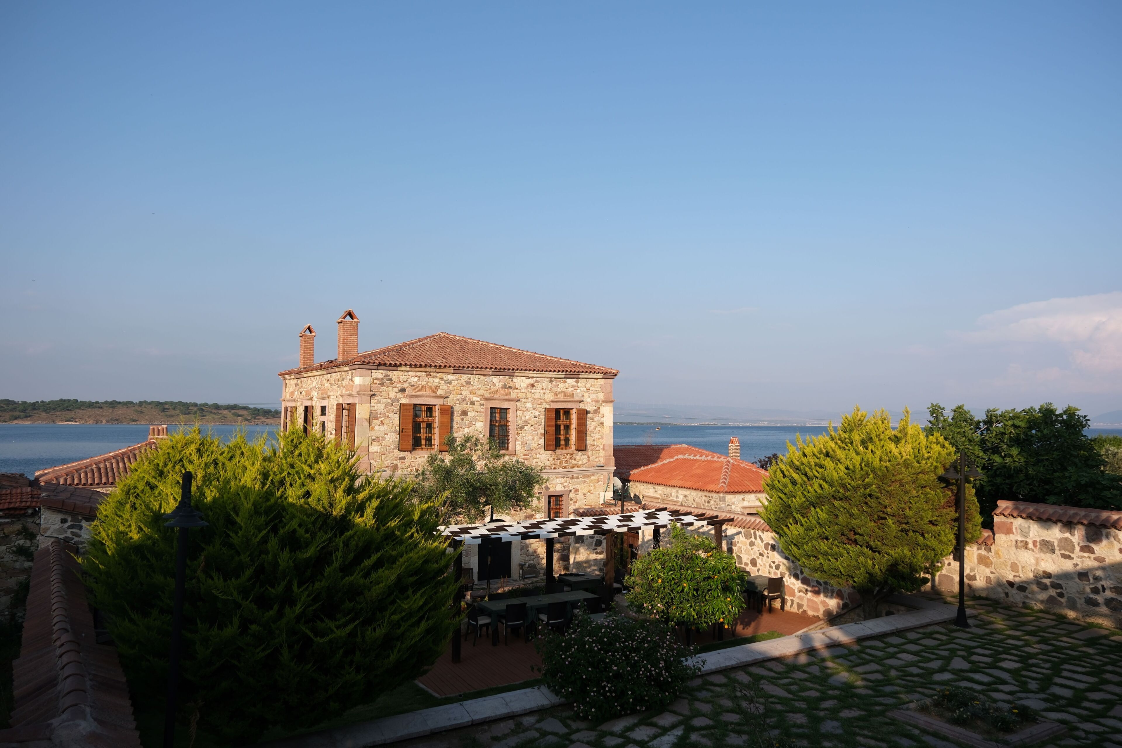 A stone building with a tiled roof, surrounded by greenery and a view of the sea.