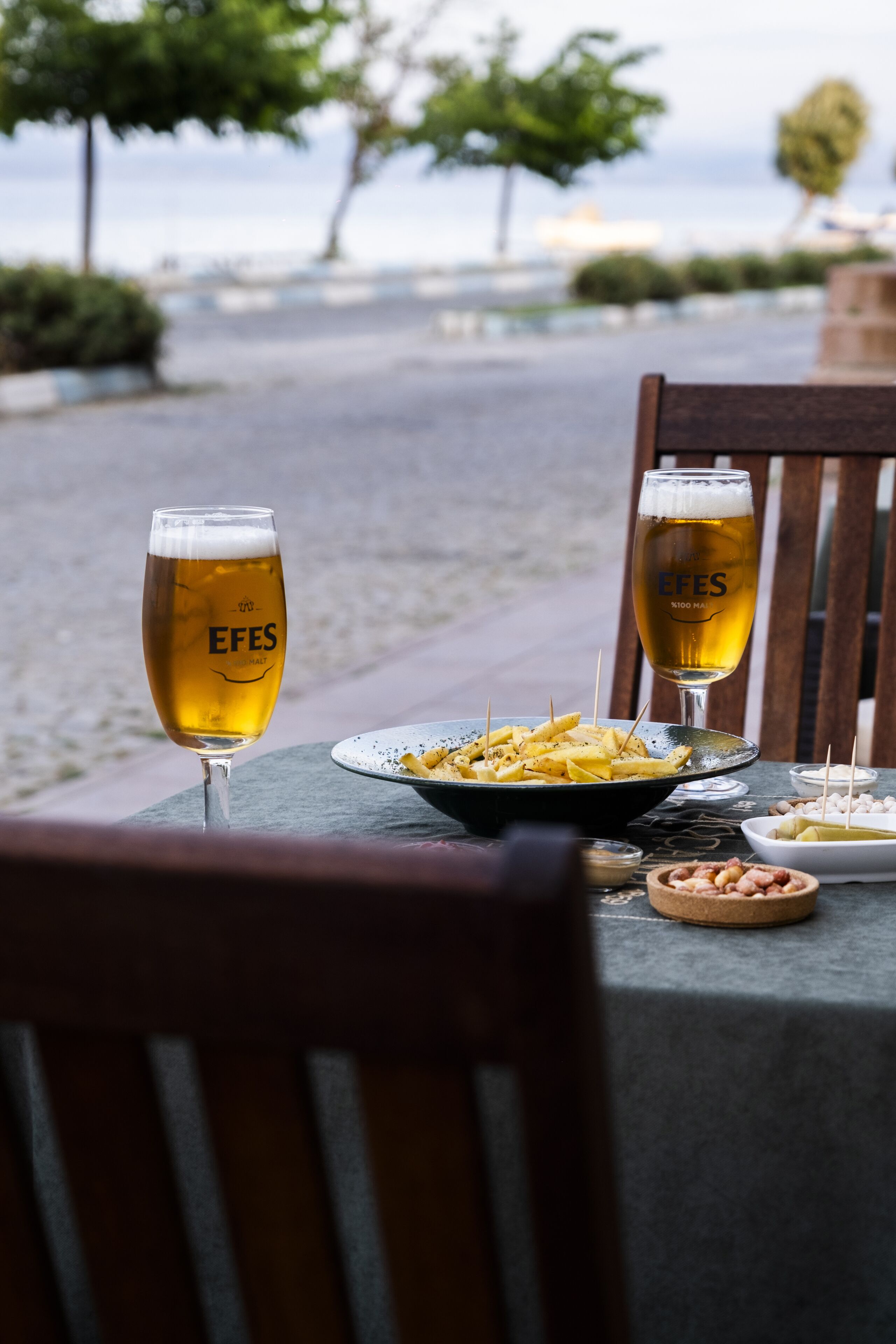A table with two glasses of beer, a plate of food, and a wooden chair.