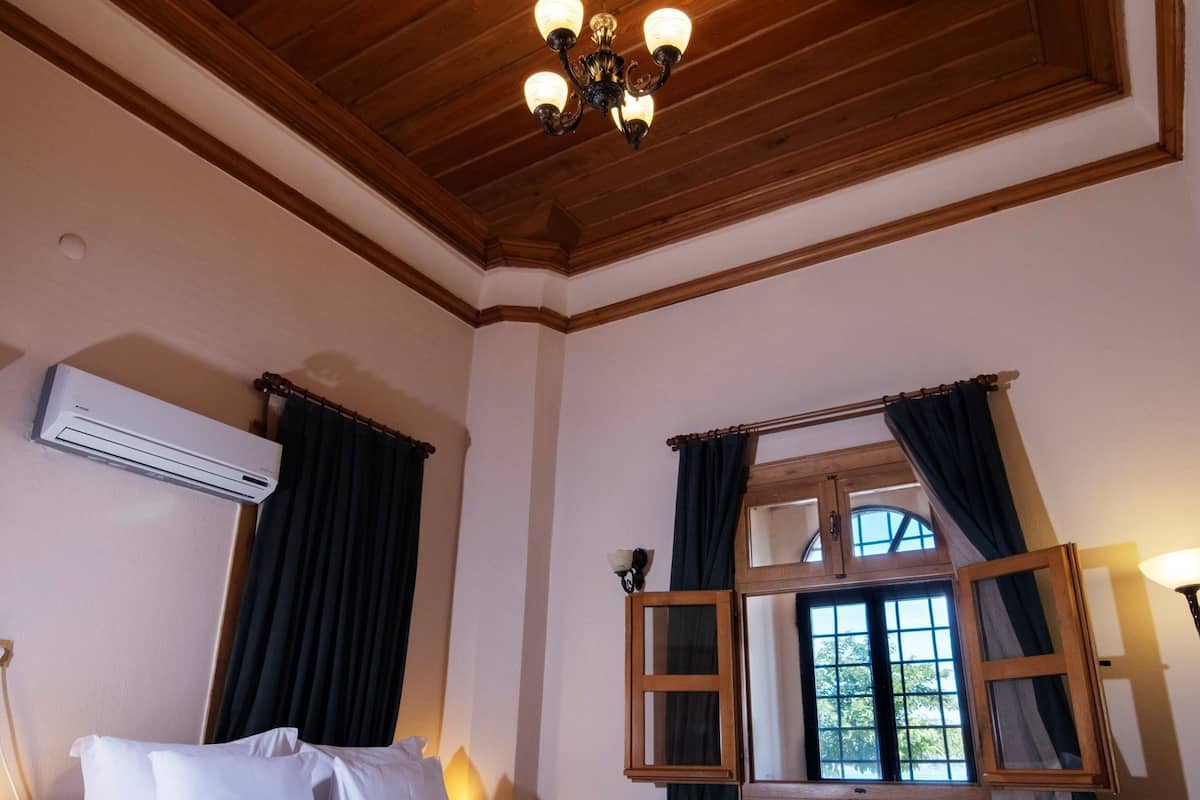 Un dormitorio con techo de madera, una cama con ropa blanca, una ventana con cortinas y una mesita de noche con lámpara.