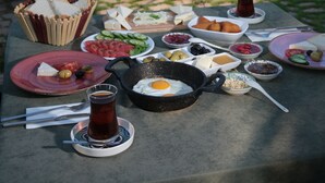 Una mesa servida con alimentos para el desayuno, como huevos, pan y una bebida, frente a un edificio de piedra con vista al mar.