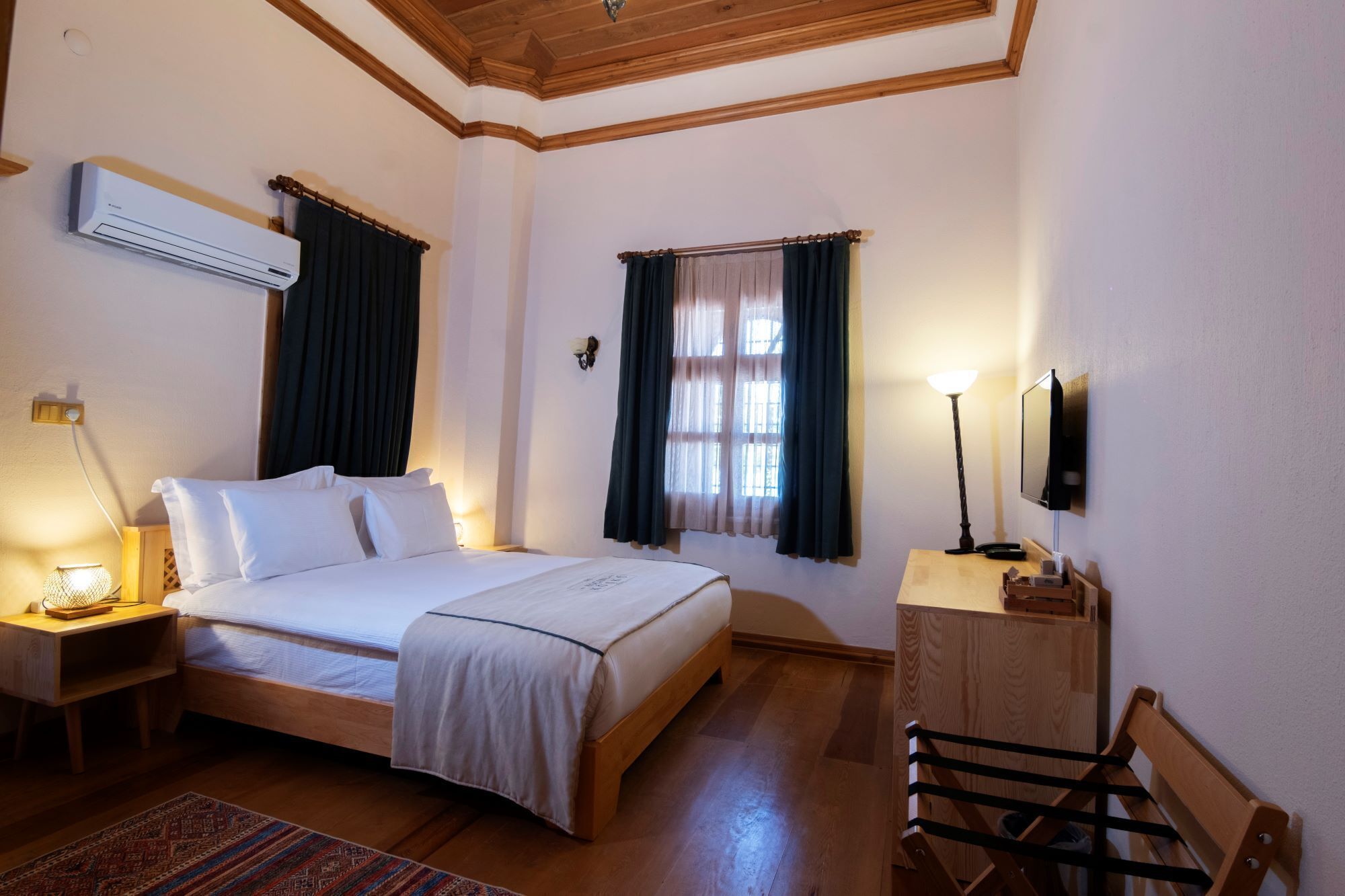 A hotel room with a bed, bedside table with lamp, air conditioning unit, window with curtains, and a TV mounted on the wall.