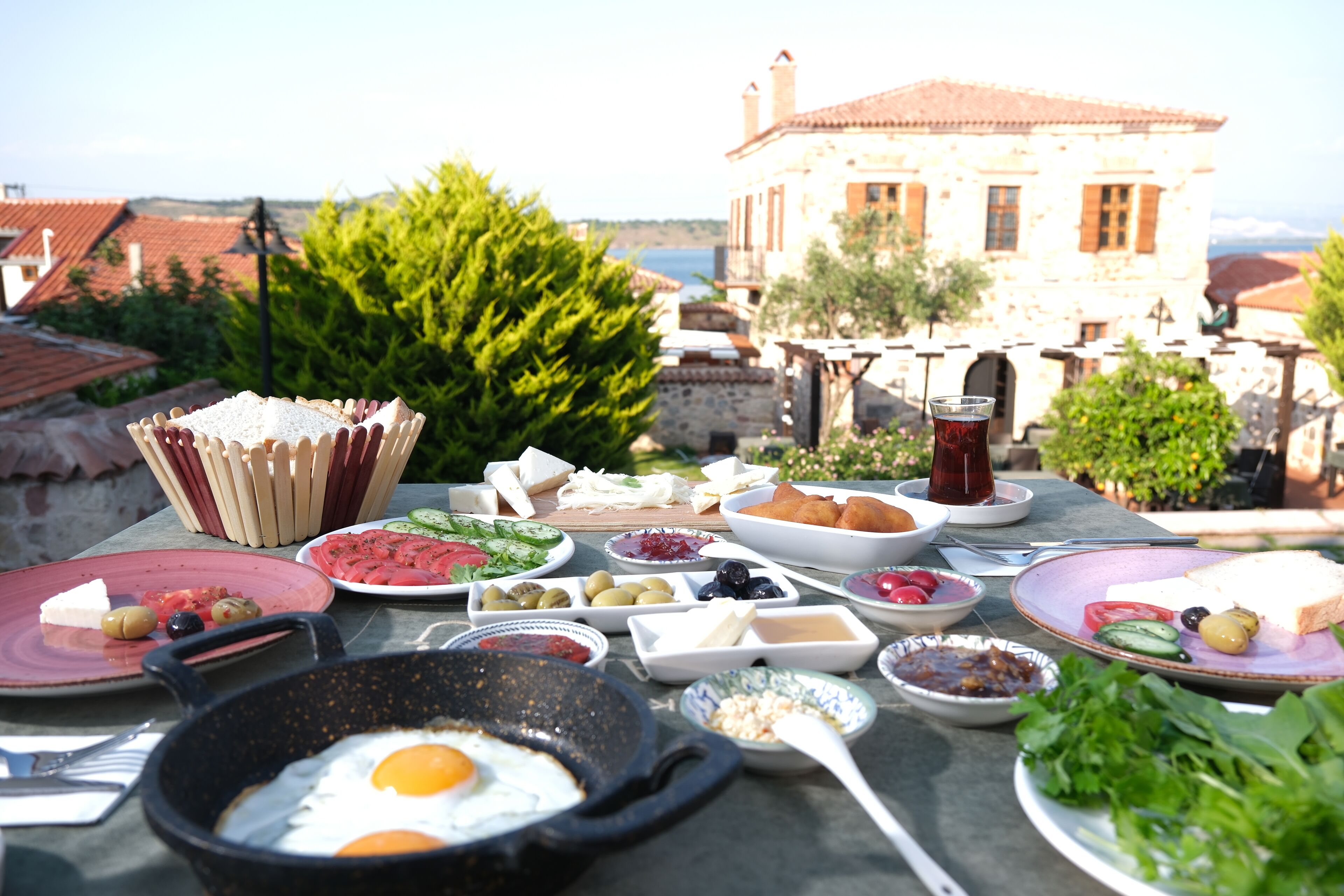 A table set with various breakfast items including eggs, bread, olives, and vegetables, with a view of a building and landscape in the background.