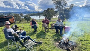 Un groupe de personnes est assis sur des chaises de jardin autour d’un feu de camp dans un champ herbeux, avec un lac et des montagnes en arrière-plan.