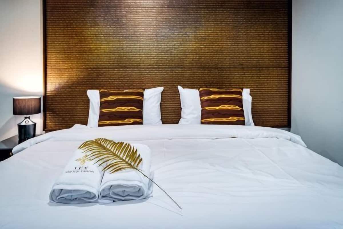 Ein ordentlich bezogenes Bett mit weißen Leinen und einem gefalteten Handtuch, ein hölzerner Kopfteil und eine Nachttischlampe.