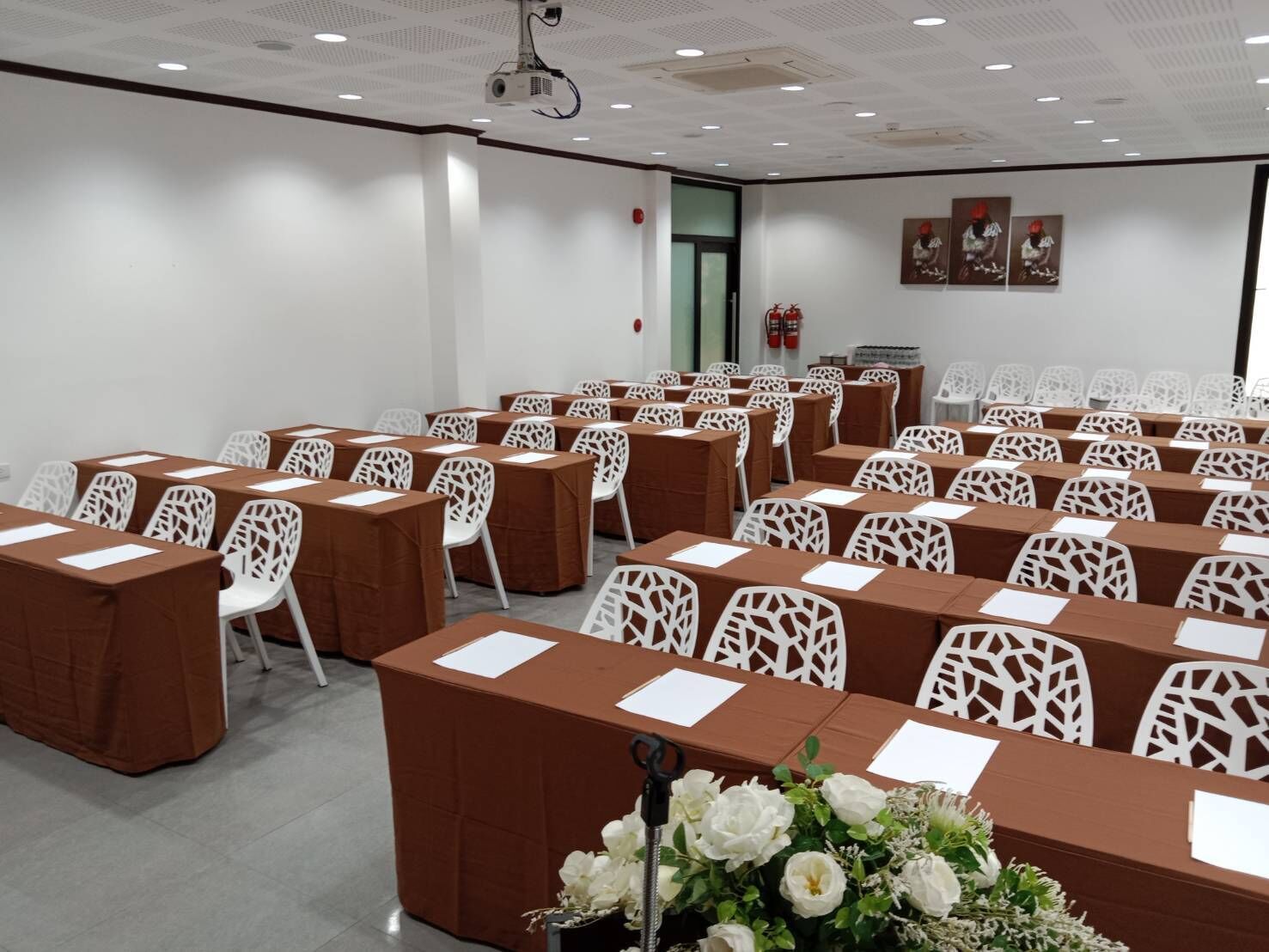 A conference room with rows of tables and chairs, a projector on the ceiling, and a fire extinguisher on the wall.