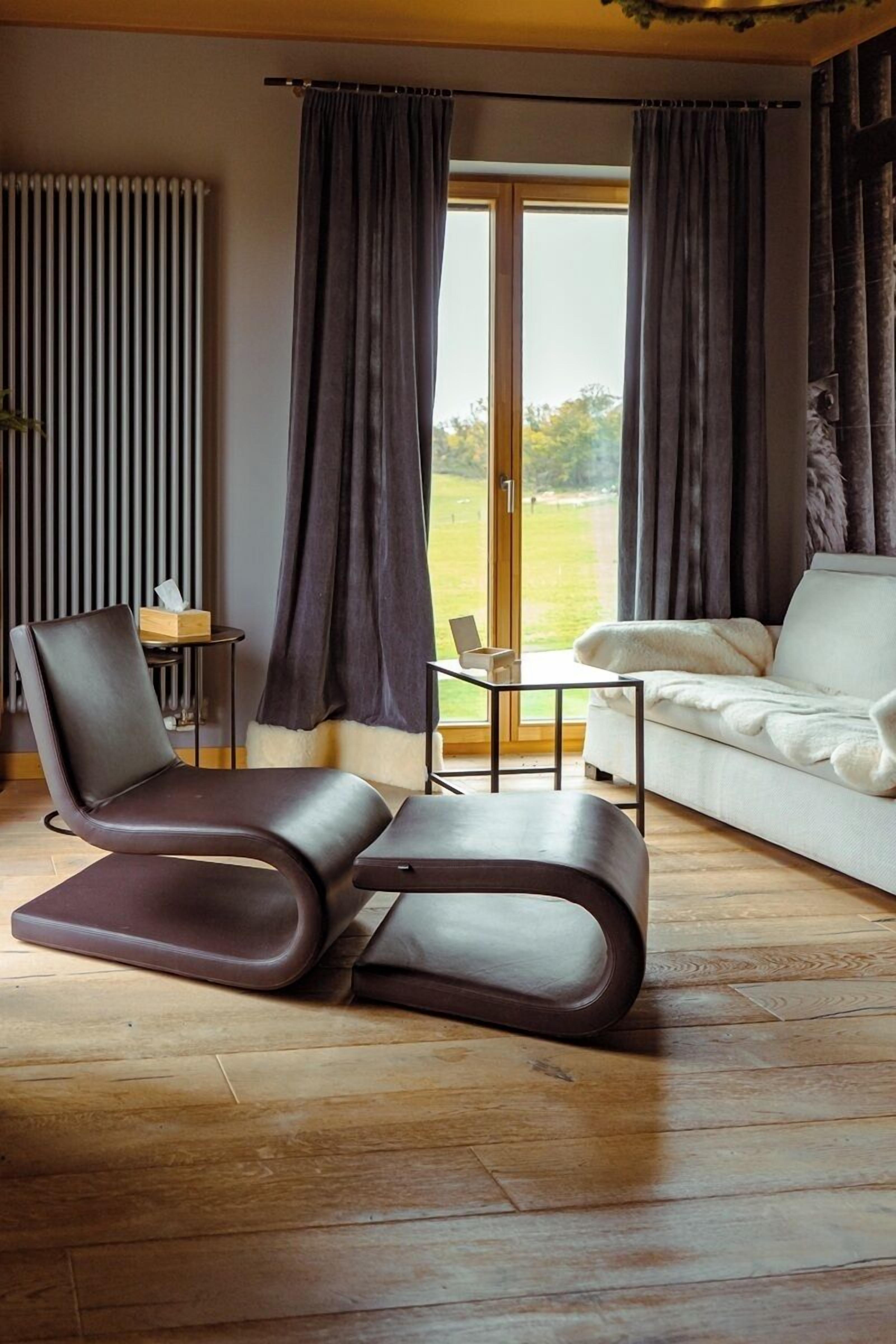 En moderne stue med sofa, lænestol, lille bord og udsigt til det fri gennem store vinduer.