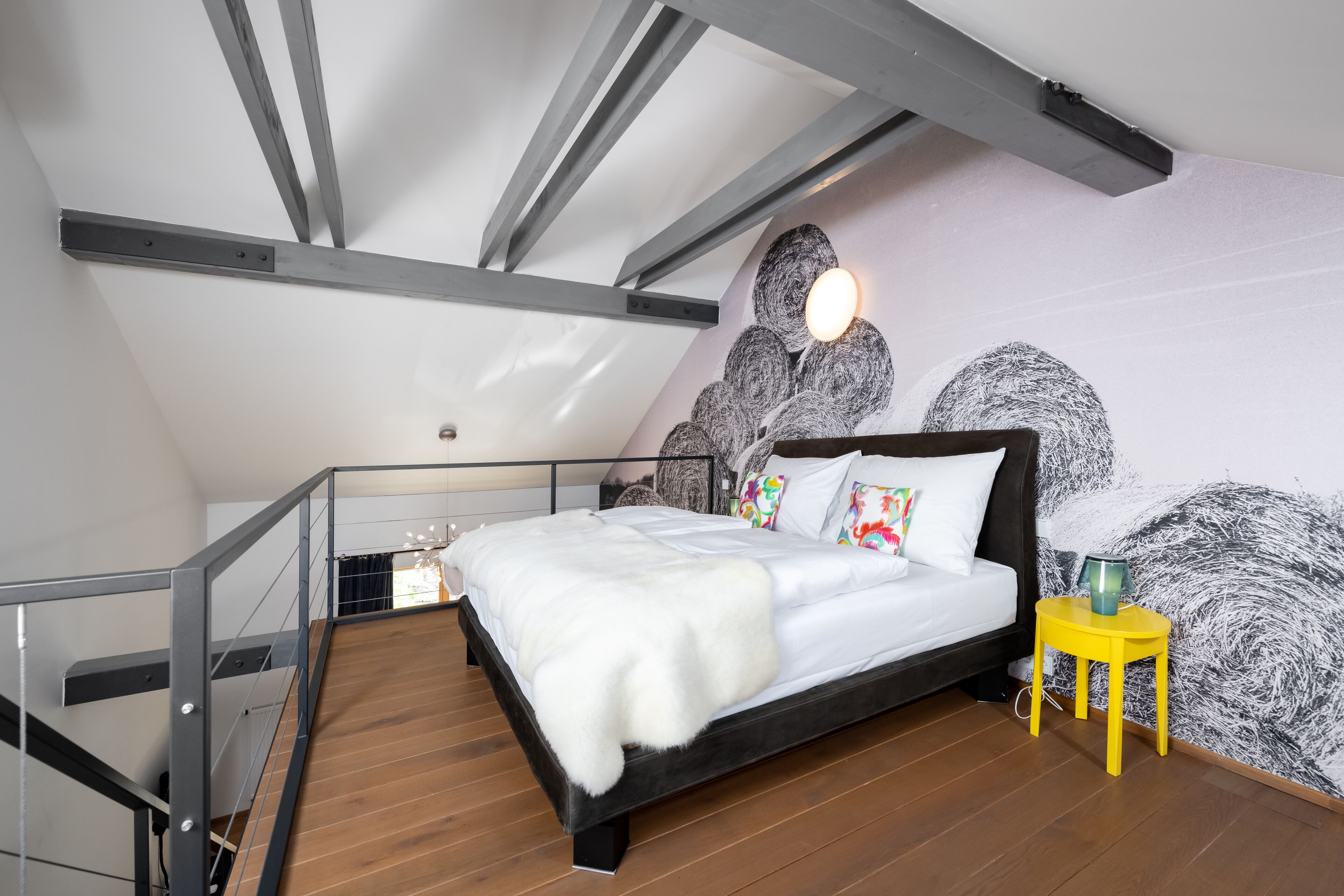 A bedroom with a sloped ceiling, a wooden floor, a bed with white bedding, a yellow side table, and a wall with a textured mural.