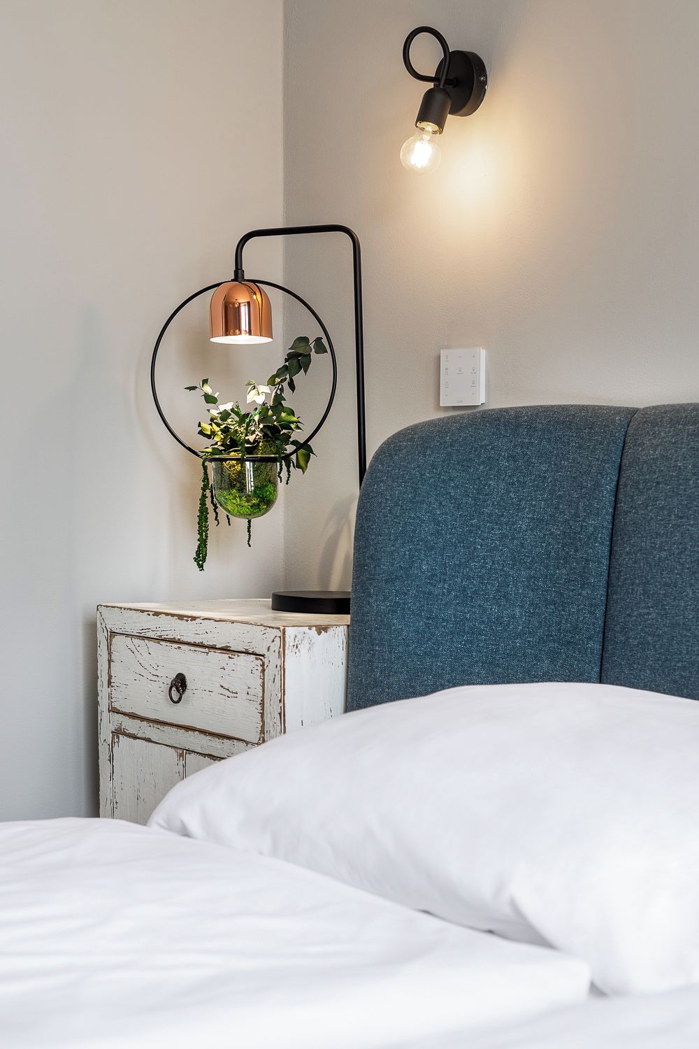 A bedroom with a bed, a white nightstand, a hanging plant, and a wall-mounted light fixture.