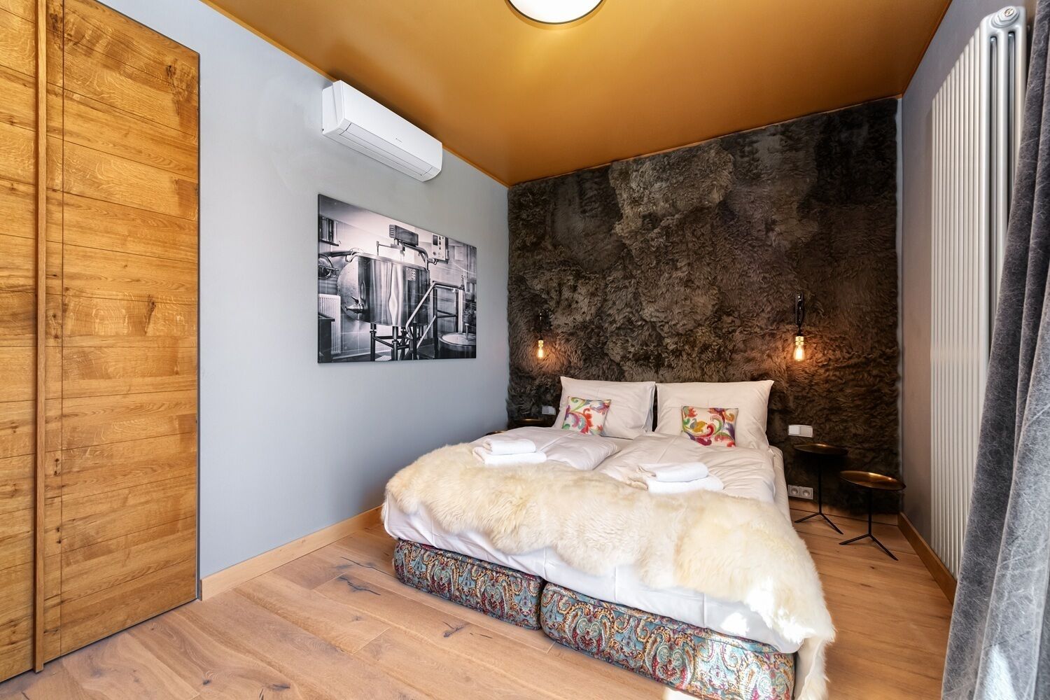 A bedroom with a wooden wardrobe, a bed with white bedding, a textured wall, and a small table with a lamp.