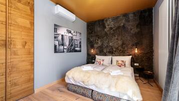 A bedroom with a wooden wardrobe, a bed with white bedding, a textured wall, and a small table with a lamp.