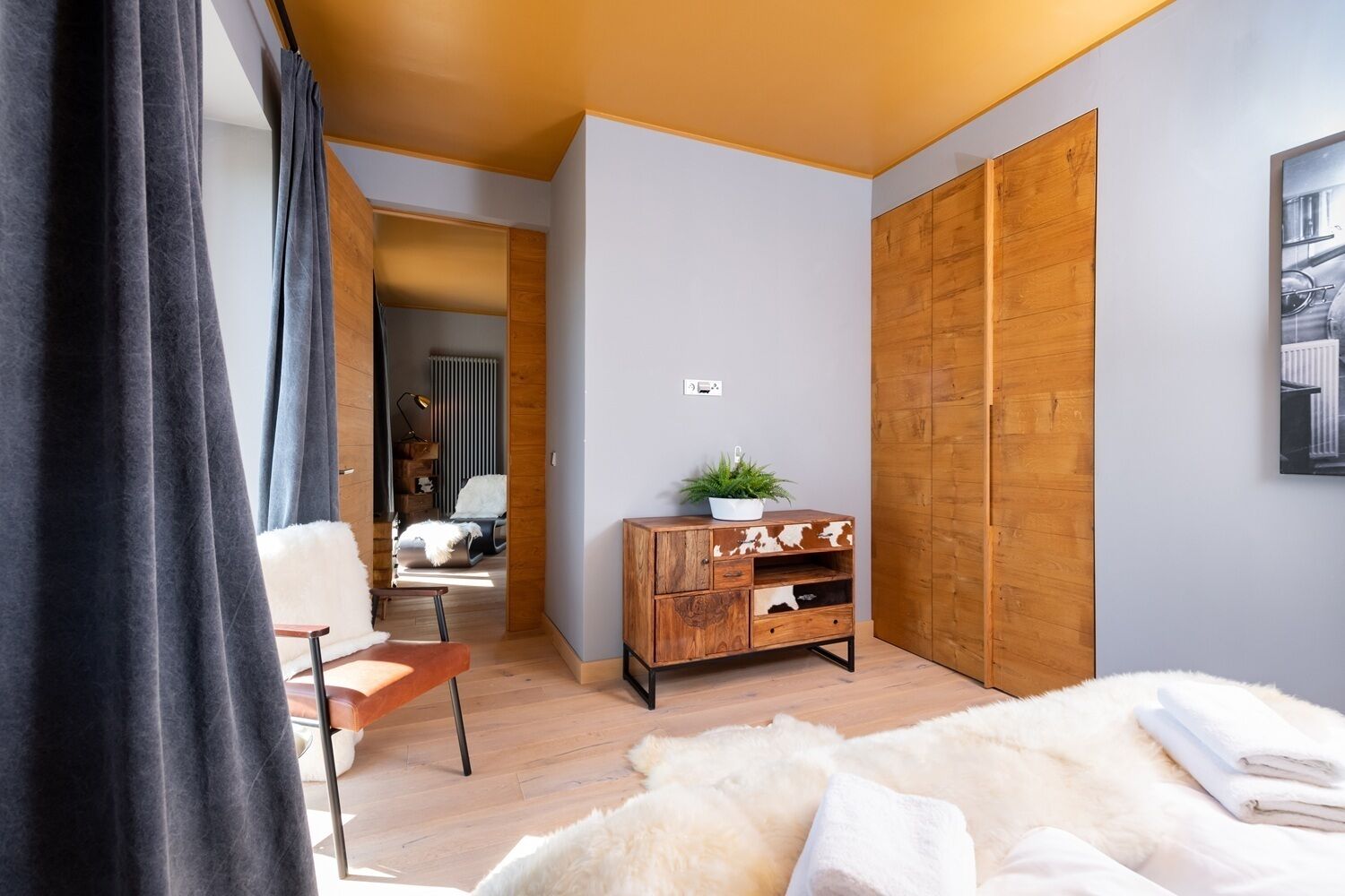 Un dormitorio moderno con un armario de madera, una cómoda pequeña de madera, una alfombra blanca mullida y una cama con ropa de cama blanca.