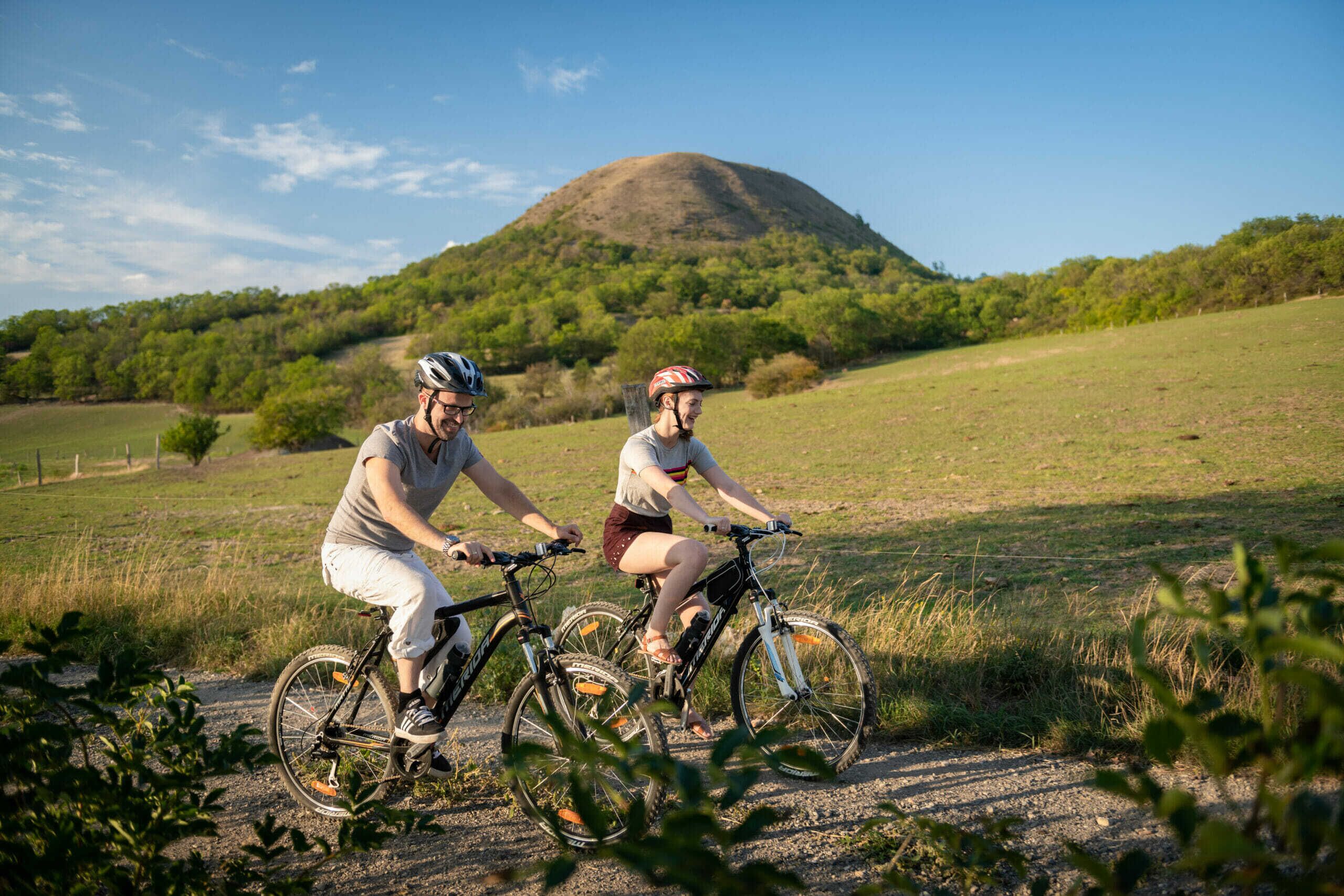 Two cyclists on a dirt path with a hill and trees in the background.