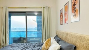 A bedroom with a bed, a view of the city through a large window, and three framed artworks on the wall.