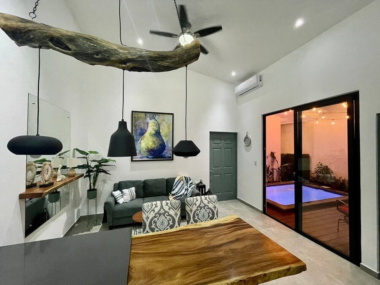 A modern living room with a wooden coffee table, a sofa, a dining table, pendant lights, a ceiling fan, and a sliding glass door leading to a pool area.