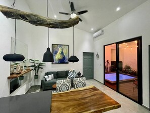 A modern living room with a wooden coffee table, a sofa, a dining table, pendant lights, a ceiling fan, and a sliding glass door leading to a pool area. - House with private pool and fully equipped BBQ (Jaco)