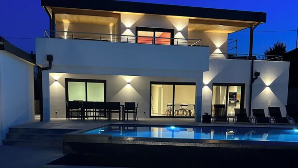 Una casa moderna de dos pisos con piscina, mobiliario exterior y una entrada bien iluminada.