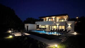 Una casa moderna de dos pisos con piscina, mobiliario exterior e iluminación ambiental.