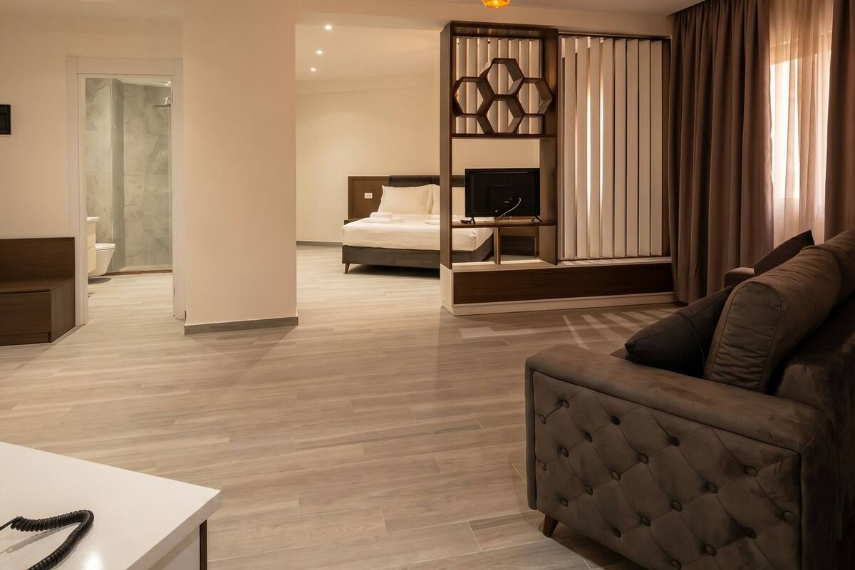 A modern hotel room with a sofa, a TV, a wooden partition, and a bathroom visible in the background.