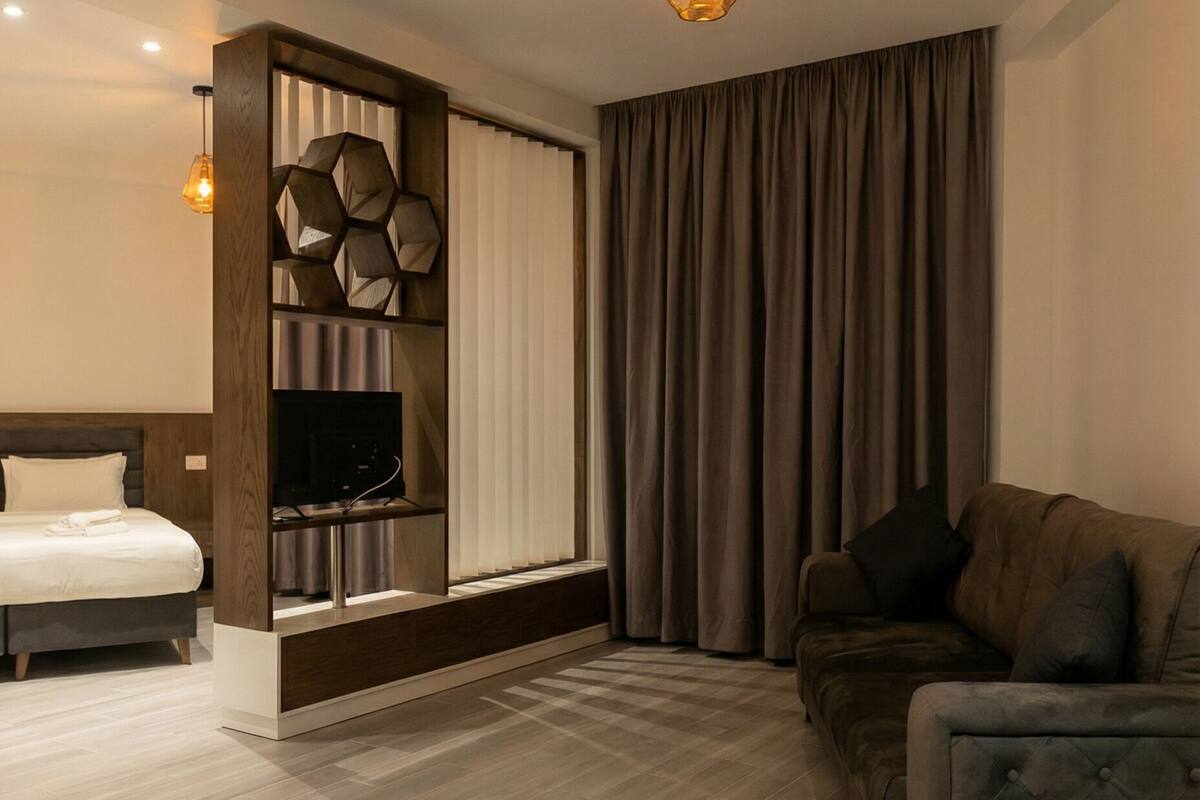 A modern living room with a sofa, a TV on a wooden stand, a bed, and a large window with curtains.