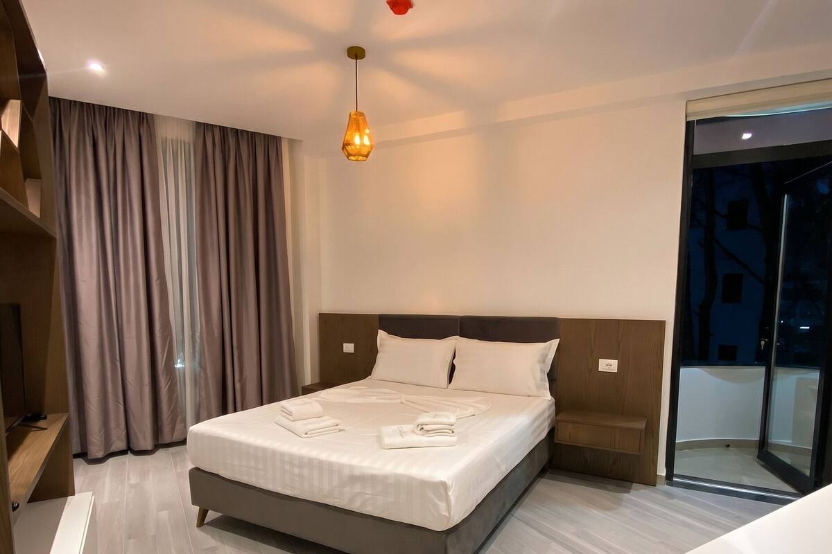 A modern hotel room with a large bed, a bedside table, a wardrobe, a sliding glass door, and a pendant light.
