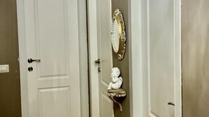 Un pasillo con dos puertas blancas, una araña, un espejo con marco dorado y un busto en una estantería.