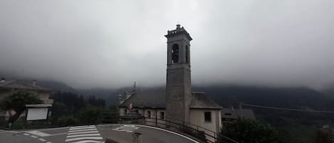 A church with a bell tower, surrounded by a hilly landscape and a cloudy sky.