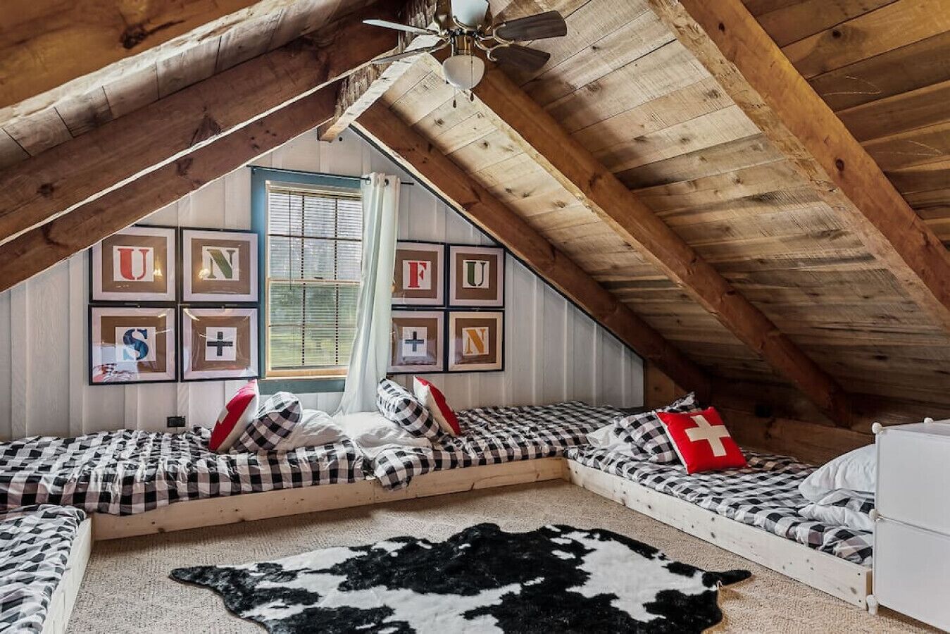 Ett mysigt sovrum på vinden med ett sluttande tak, synliga träbjälkar, en takfläkt och ett fönster med persienner.