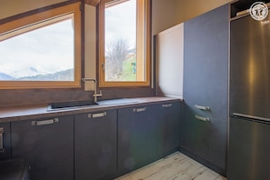 A modern kitchen with dark cabinets, a stainless steel refrigerator, and a window with a view of a green landscape. - Chalet Panoramic - Valloire (VALLOIRE)