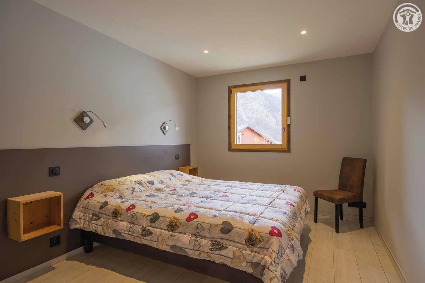 A bedroom with a bed, a window, a chair, and a small wooden shelf.
