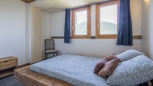 Un dormitorio con cama, dos ventanas con cortinas, una silla y una mesita de noche de madera.