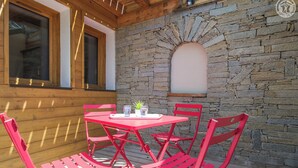 Una terraza de madera con un muro de piedra, una mesa roja con sillas y un ventanal con persianas.