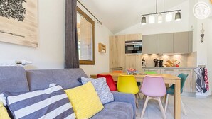 Un salon moderne avec un canapé gris, des fauteuils colorés, une table à manger en bois et une cuisine en arrière-plan.