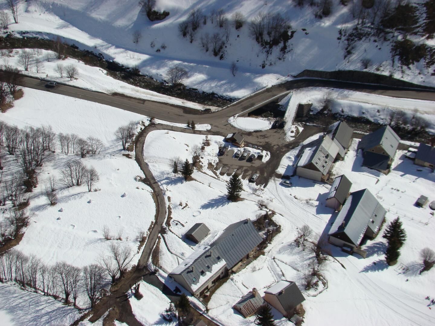 An aerial view of a snow-covered residential area with houses, a winding road, and a small parking lot.