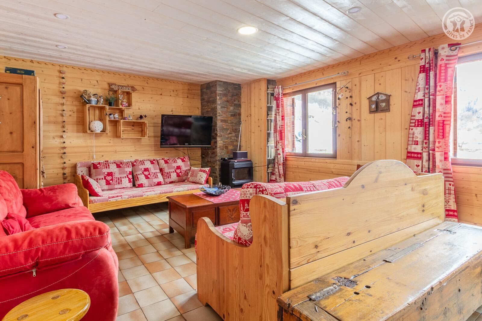 Ein Interieur einer Holzhütte mit einer Couch, einem roten Sessel, einer Holzbank, einem Fernseher und einem Kamin.