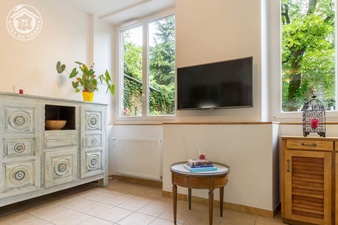 A room with a white cabinet, a television mounted on the wall, a small round table with books, and a window with a view of greenery.