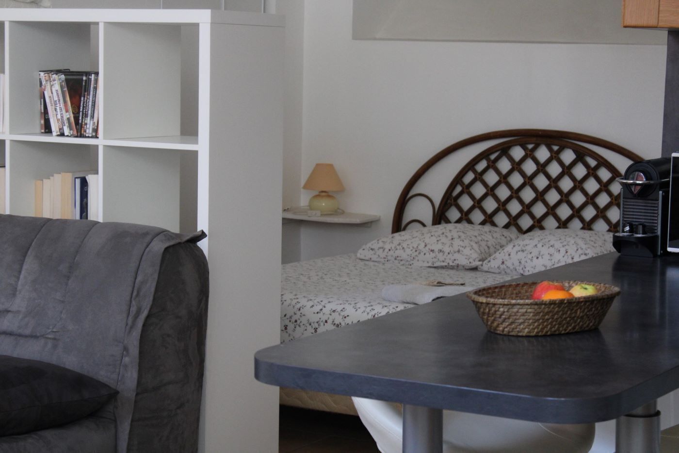 A bedroom with a bed, a bedside table with a lamp, a wicker basket with fruit, and a grey sofa.