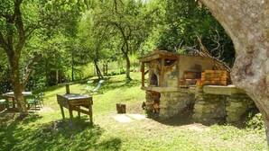 A rustic outdoor setting with a stone-built structure, a wooden table, and chairs, surrounded by lush greenery.