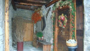 A rustic stone-walled hallway with a wooden door adorned with festive decorations. A bench and a potted plant are visible in the foreground.