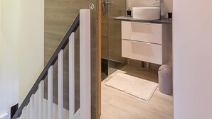 A modern bathroom with a glass shower, a white sink, and a wooden door leading to a staircase.